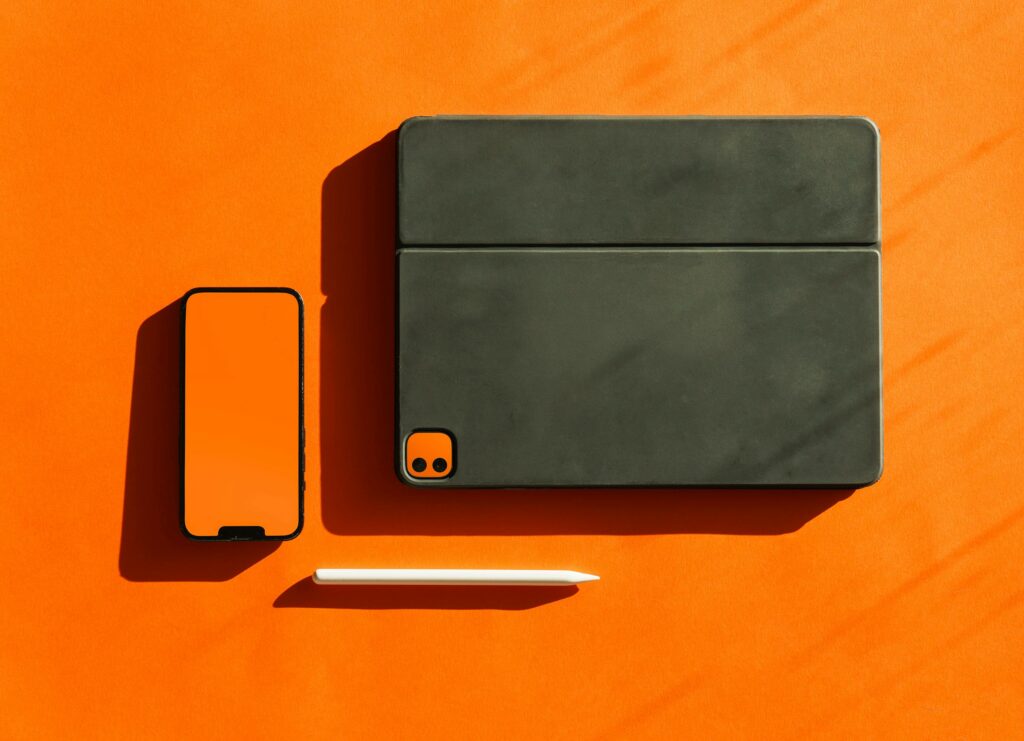 Stylish flat lay of digital devices and stylus on a vibrant orange surface, ideal for technology themes.