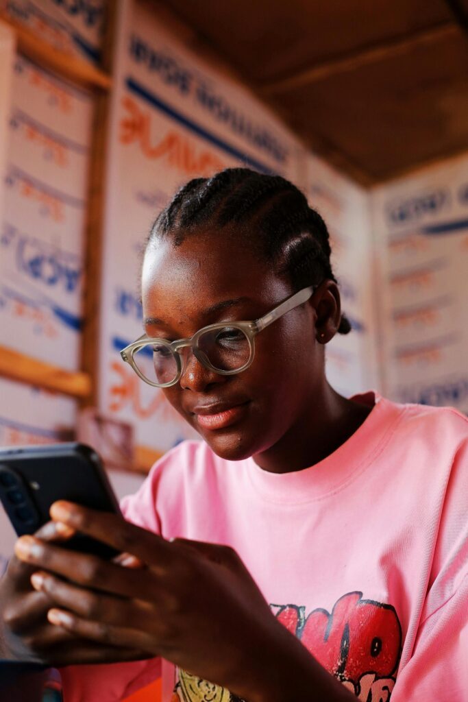 Why Tech Skills Are the New Degree in Nigeria Young girl engrossed on her smartphone indoors, wearing glasses and a casual pink shirt.