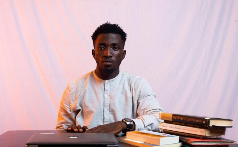An African man sitting at a desk with a laptop and books, looking serious and focused indoors.
